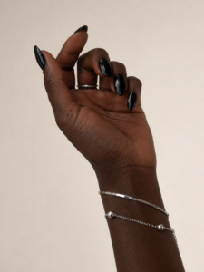 Dark-skinned hand with cool undertone wearing silver jewelry and glossy black nails against a soft beige background